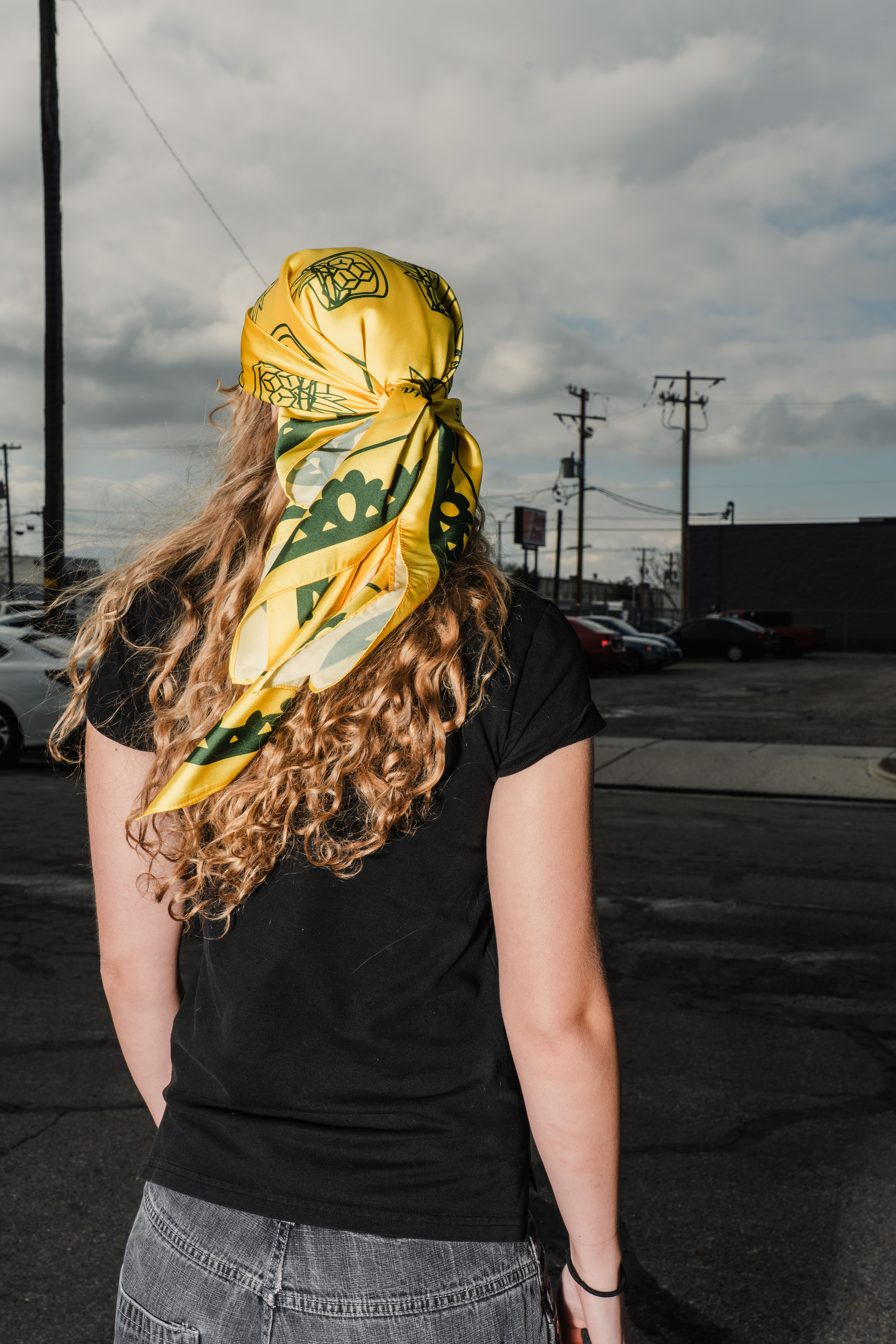 Girl with long curly hair in a black polo and jeans looking away from the camera wearing a yellow and green scarf that is branded for Buchanan's on her head.