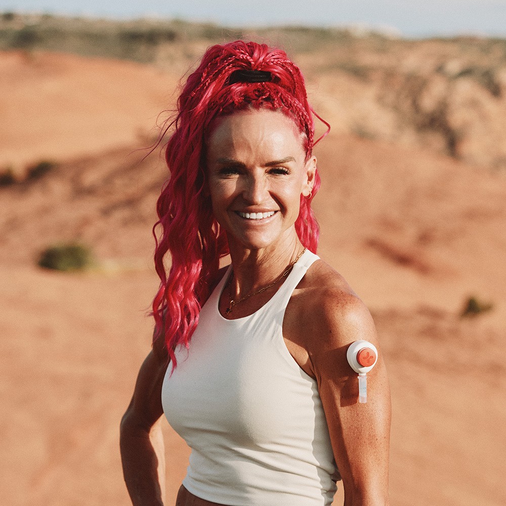 Woman outdoors in a desert landscape wearing a Rythm Health biosensor patch on her upper arm, a wearable device used for continuous health tracking and at-home biomarker monitoring.