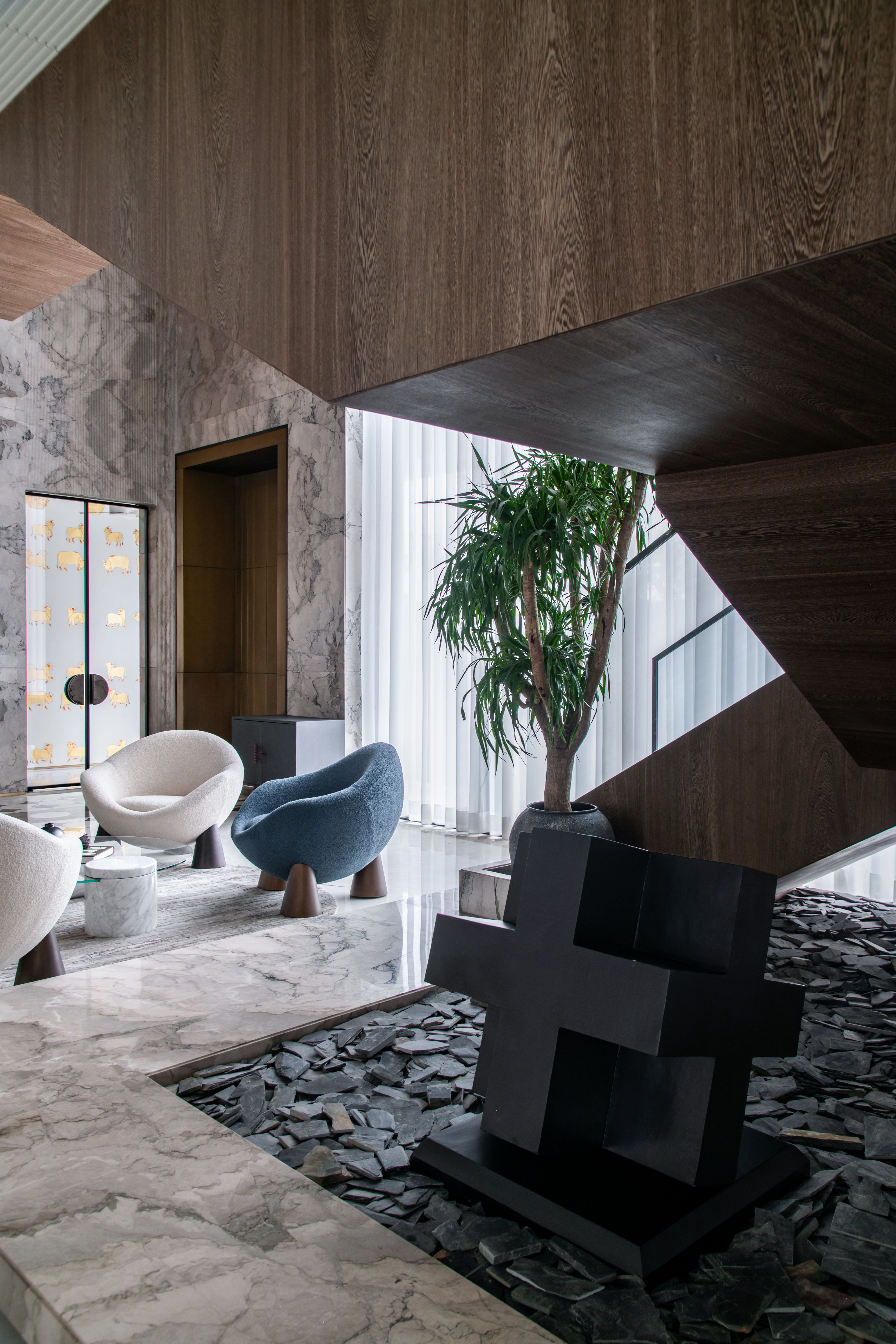 A contemporary living room featuring warm wood paneling, modern furniture, and large floor-to-ceiling windows that fill the space with natural light. The interior combines minimalist design with elegant finishes and indoor greenery.