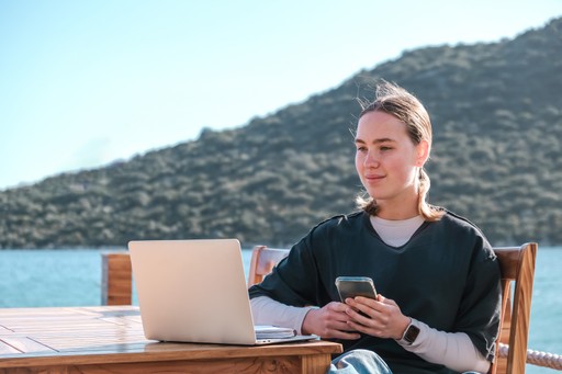 A person in scrubs sits outside by water, smiling and using a phone, with a scenic mountain background.