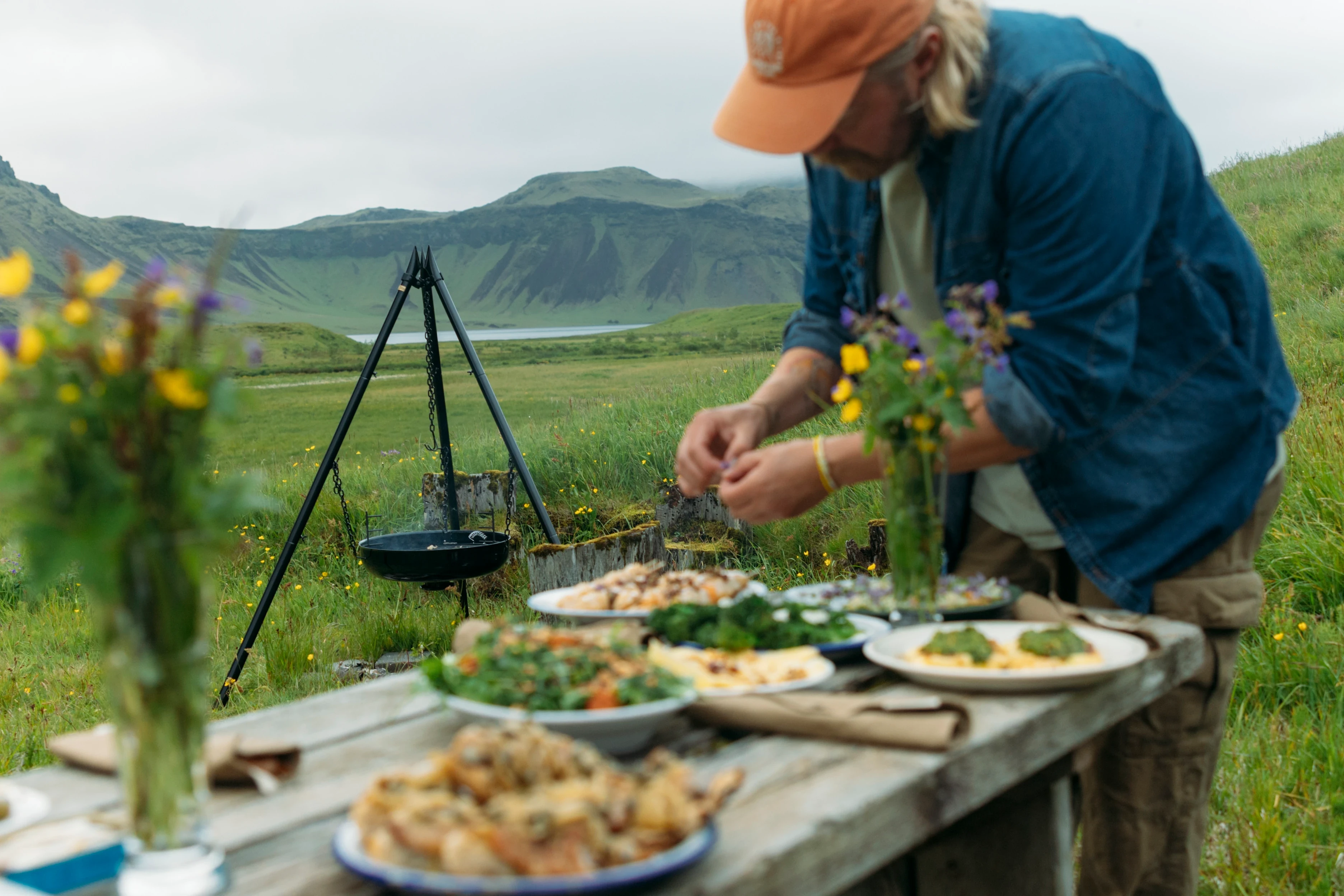 A group of four people enjoys an outdoor picnic on a rustic wooden table set amidst a lush green valley, surrounded by rolling hills under a cloudy sky, with a variety of dishes and drinks elegantly arranged on the table.