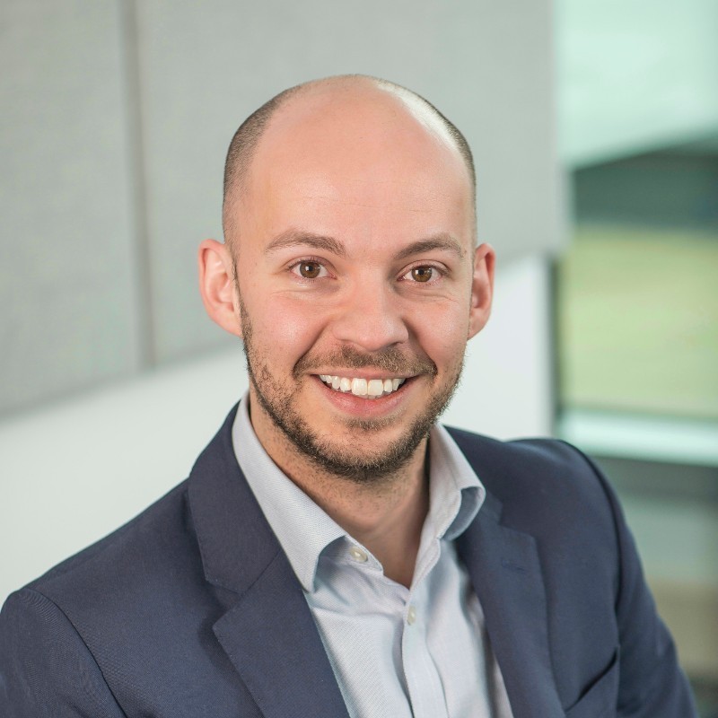 Man in glasses and a dark blazer smiling confidently, standing against a deep red background with soft sunlight streaming through.