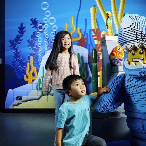 Dvoje djece smiješi se ispred LEGO izložbe s podvodnom tematikom, s raznobojnim koraljima i figurom ronioca.