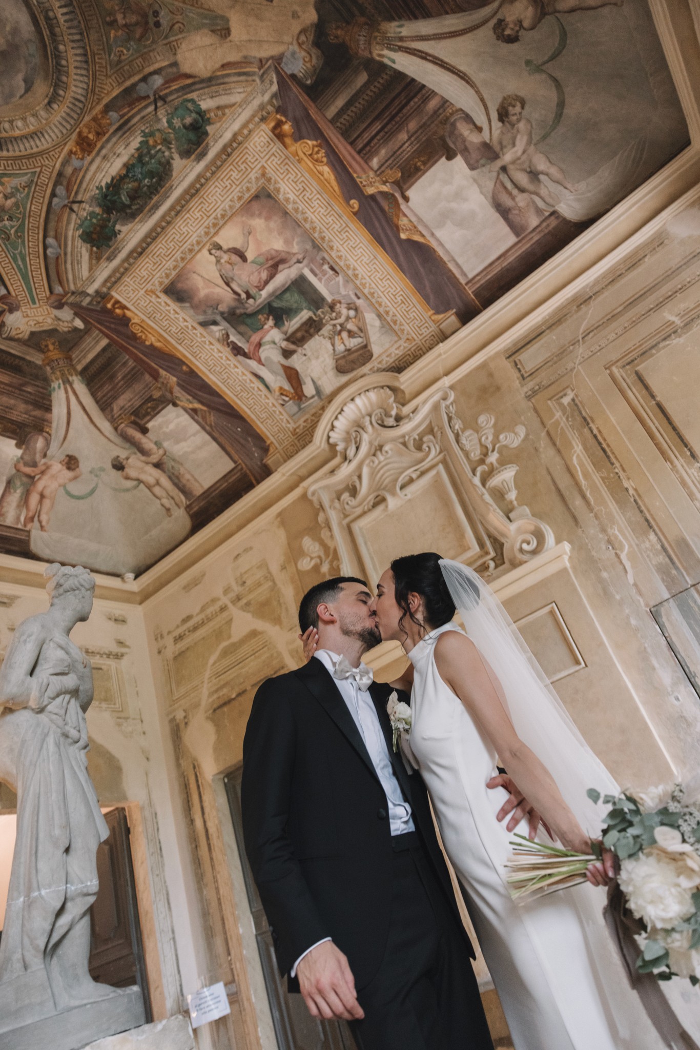 Sposi si baciano sotto gli affreschi barocchi di una sala storica, lei in abito bianco e velo, lui in smoking scuro