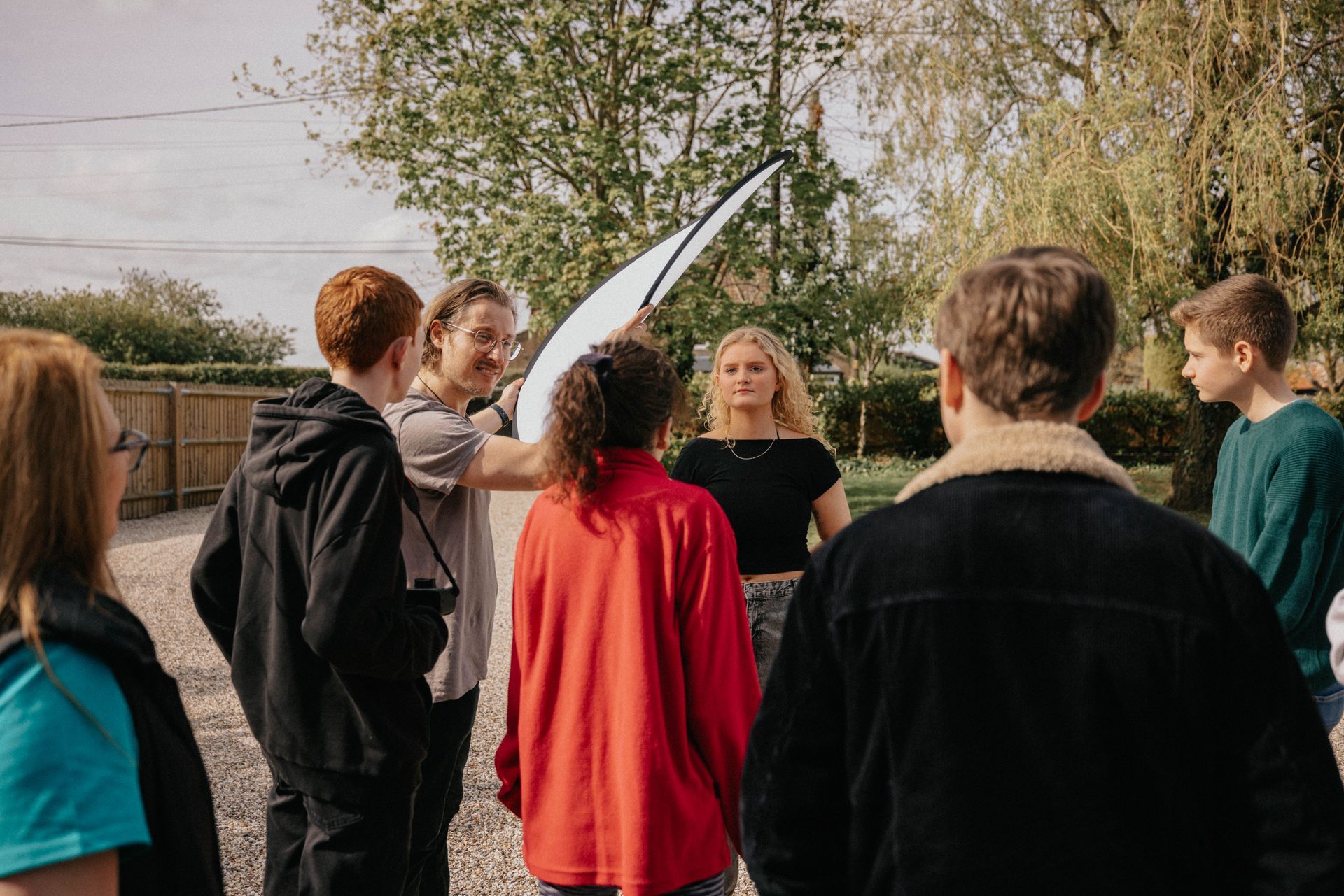 A group of people gather outdoors, listening to a speaker pointing towards a white structure in the distance.