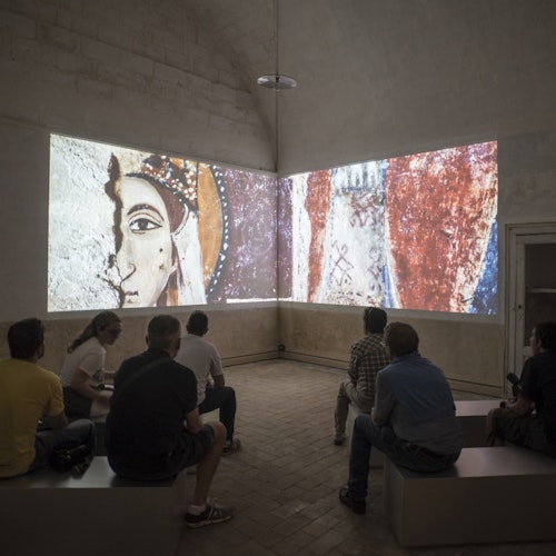 在一个灯光昏暗的房间里，人们坐在长椅上，观看投影在两面墙上的古代壁画式艺术作品。