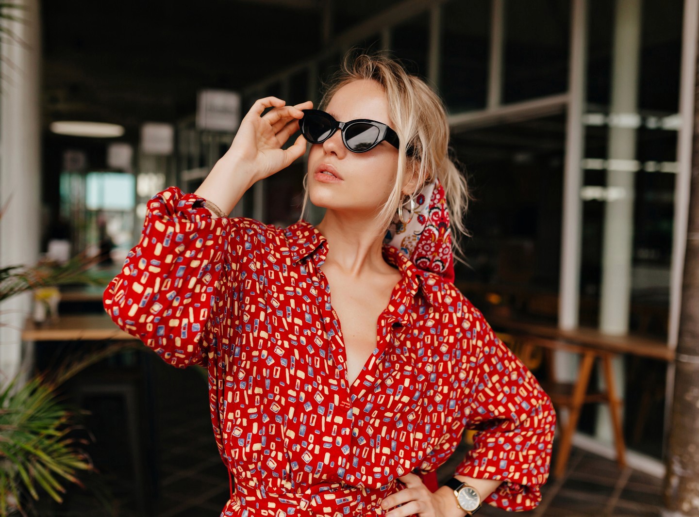 A woman in a stylish red patterned dress confidently adjusts her black sunglasses while standing outside a modern café, her hair tied back with a colorful scarf, embodying chic fashion and elegance.