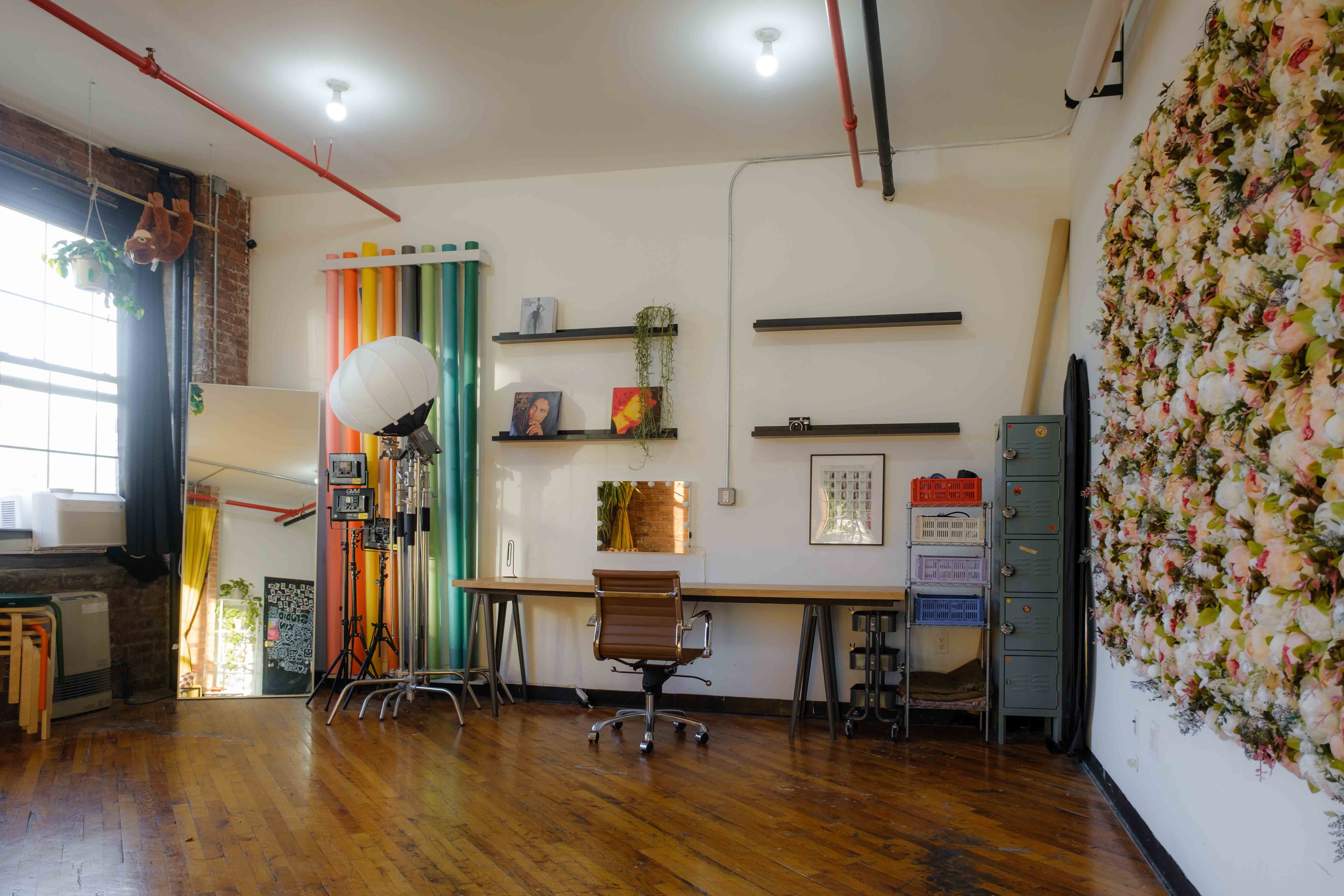 A daylight photography studio featuring a large wall of colorful backdrops, professional lighting equipment, and a workspace with shelves displaying decor and camera gear. A floral wall installation and a large mirror add artistic elements to the studio’s bright, versatile environment.