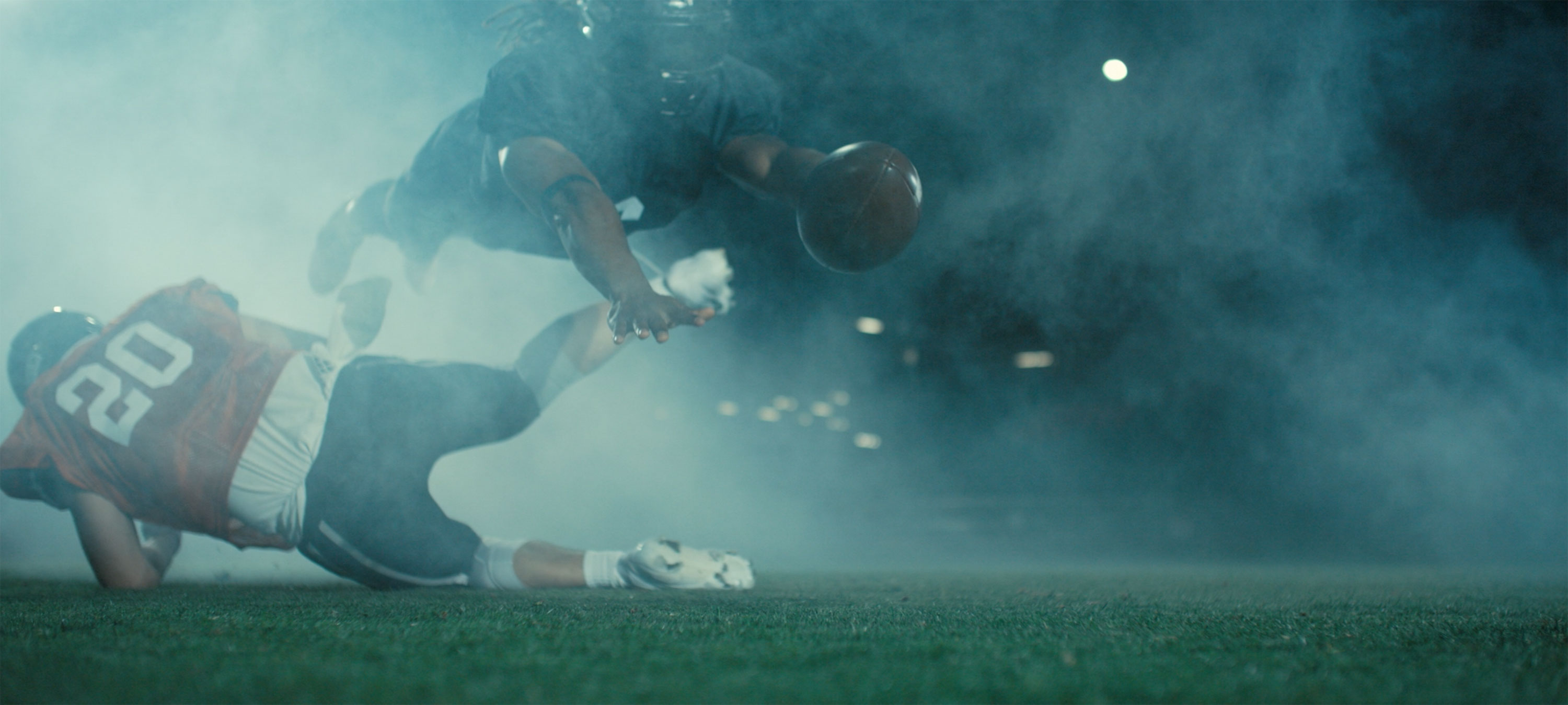 Low-angle shot of a smoky tackle: one player airborne with the ball as another falls to the turf at night.