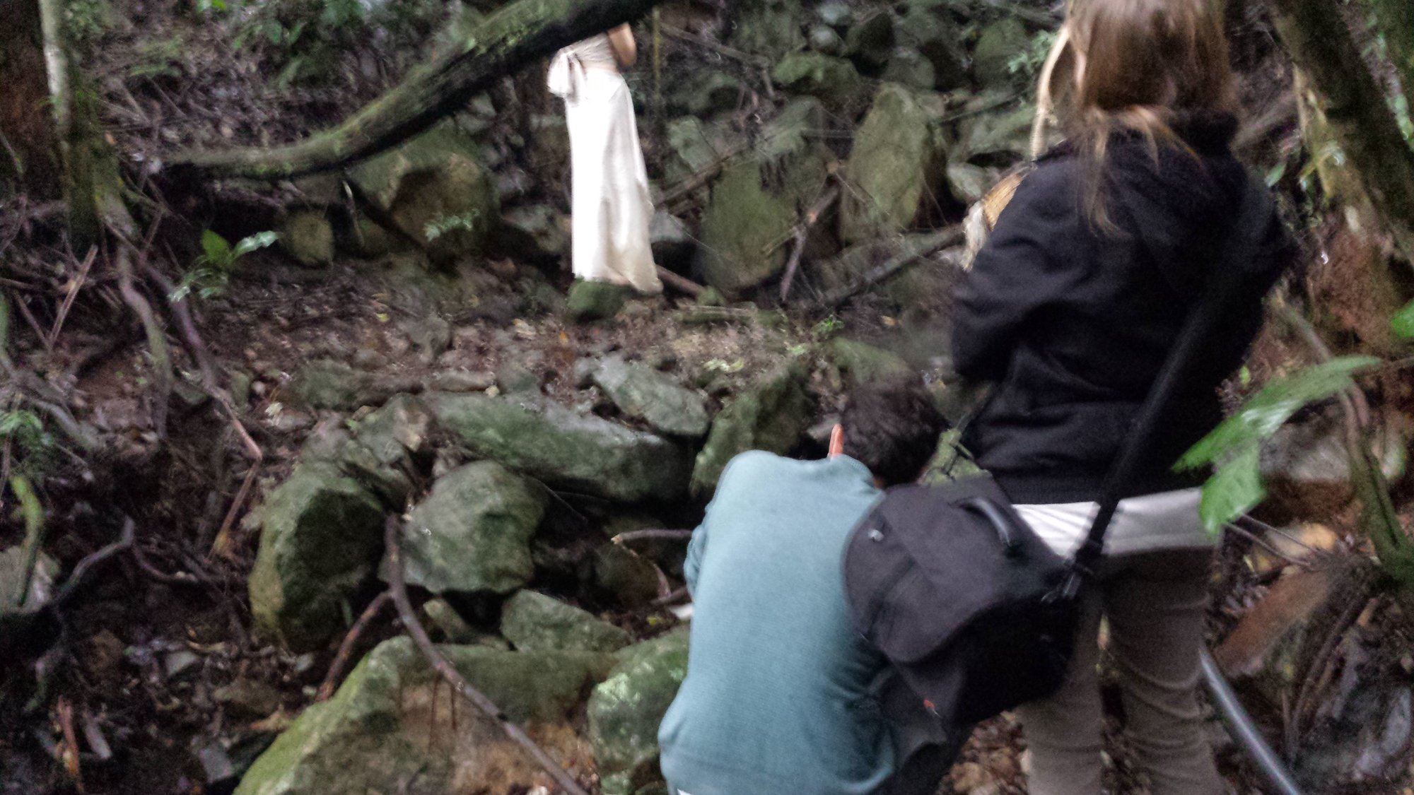 Equipe desce entre pedras da mata enquanto atriz de vestido branco aguarda, making of com Gabriela Castello