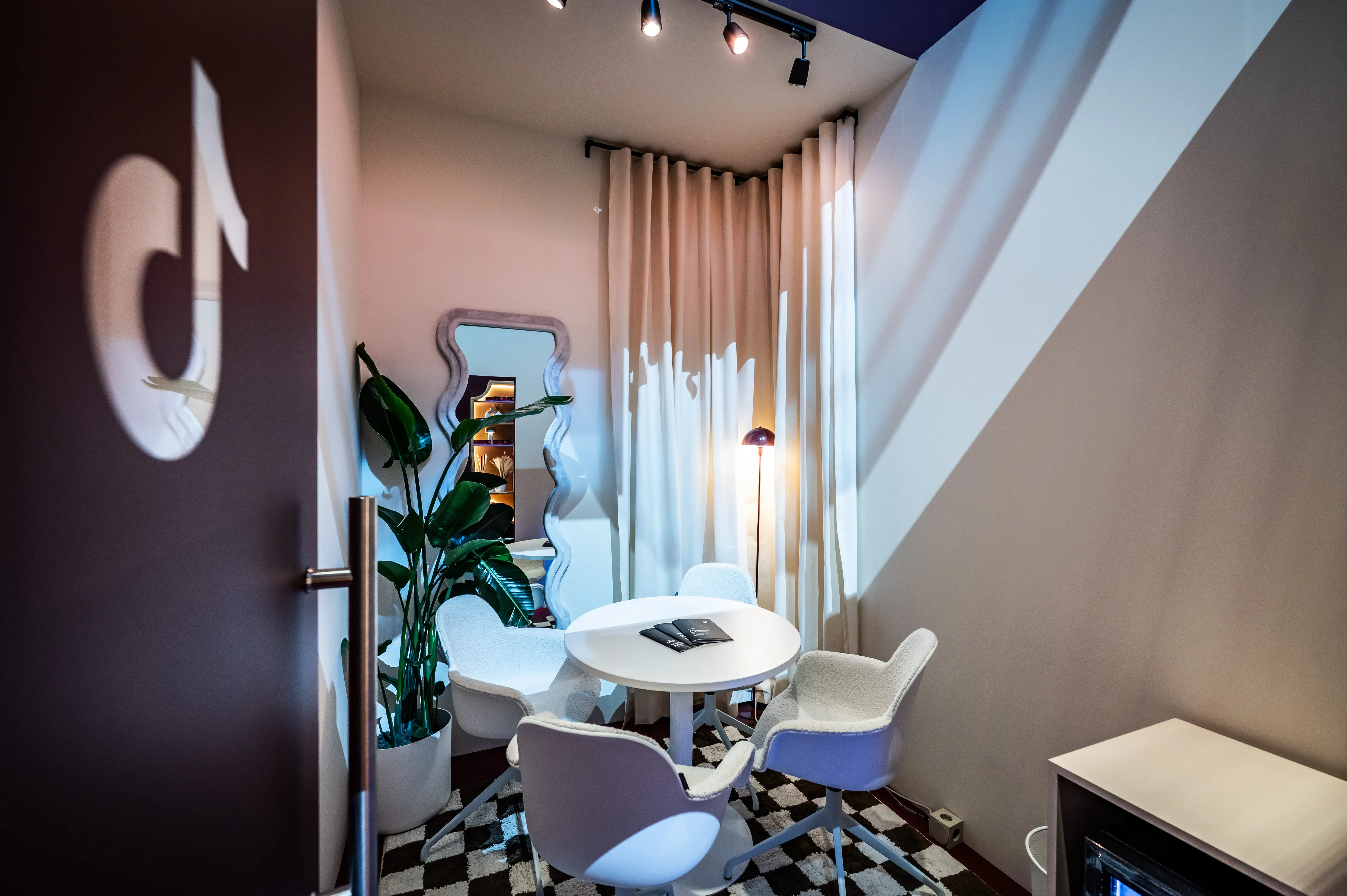 Small meeting room with white table, swivel chairs, wavy mirror, plant and checkered floor.