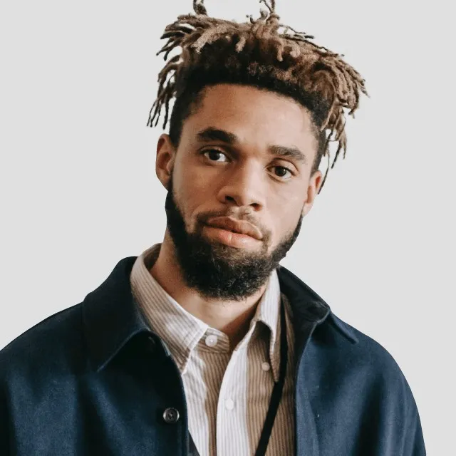 Young man with dreadlocks and beard wearing a blue jacket and striped collared shirt.