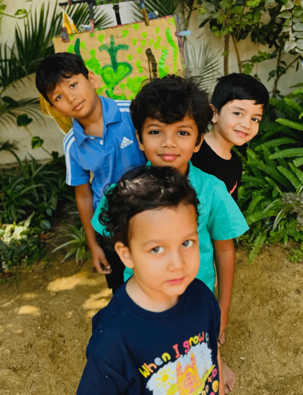 Four children pose smiling at the Nebula Montessori garden, surrounded by greenery, with a colorful sign in the background.