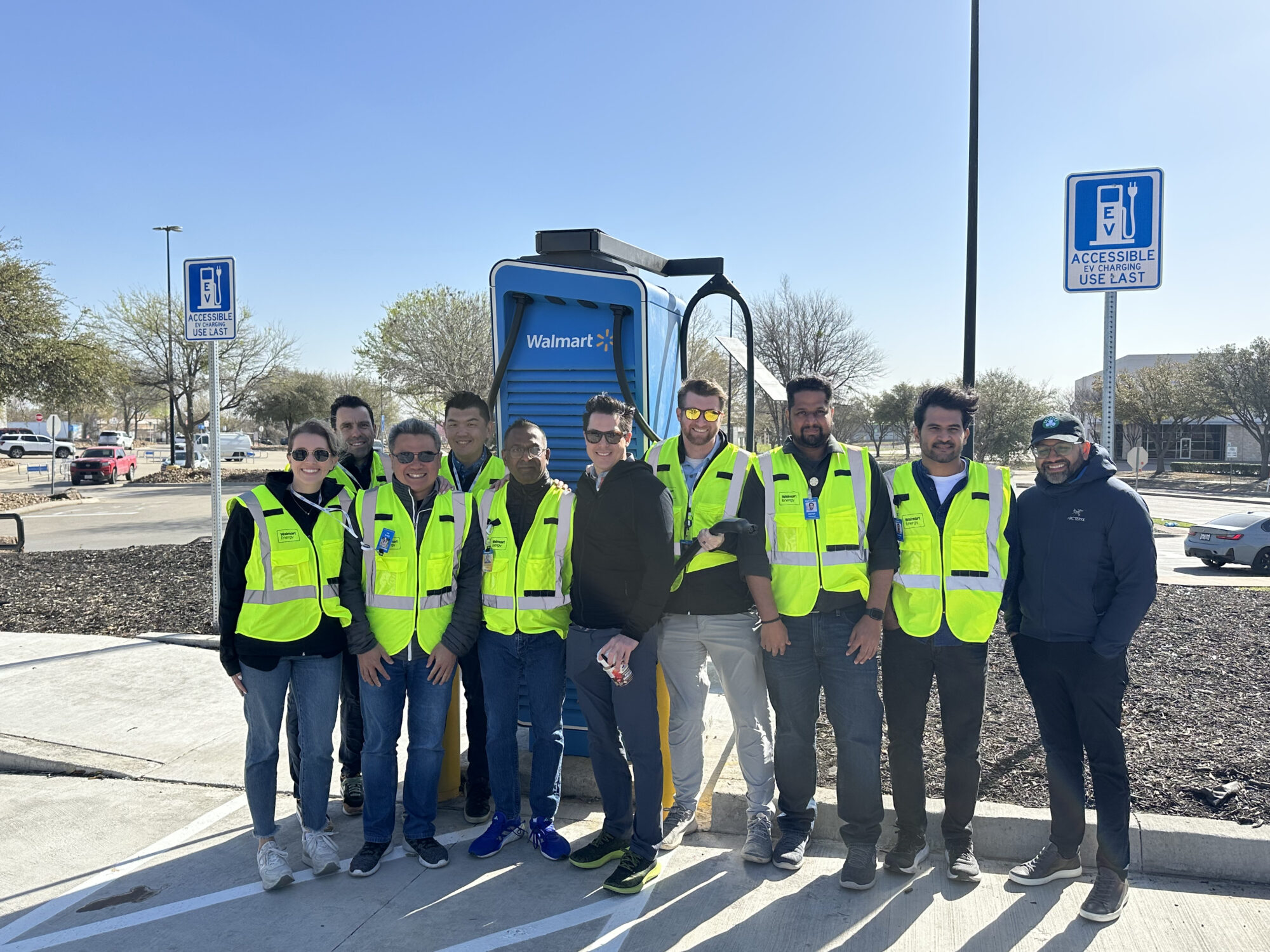 A group of people in safety vests stands together in front of a blue sign on a sunny day.