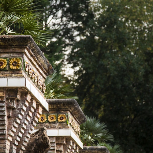 Estructura de ladrillo y piedra con azulejos decorativos de girasol, flanqueada por hojas de palma, en un fondo de denso follaje verde.
