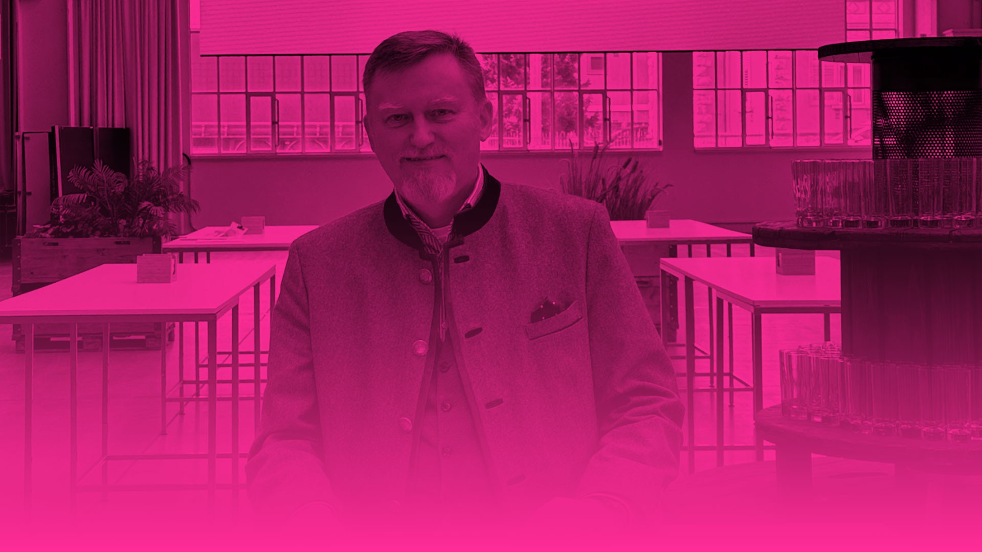 Das Foto zeigt das Porträt eines Mannes mit einem grauen Spitzbart, der leicht lächelnd in die Kamera blickt. Er trägt eine traditionelle Trachtenjacke (Janker) mit dunkler Einfassung an den Kanten und markanten Knöpfen, dazu eine Weste, ein gemustertes Hemd und ein kleines Einstecktuch. Das auffälligste Merkmal des Bildes ist ein starker, vollflächiger magentafarbener (pinker) Farbfilter, der die gesamte Szenerie einfärbt. Der untere Bildbereich verblasst zudem fließend in eine durchgehende magentafarbene Fläche. Im Hintergrund ist ein Raum zu erkennen, der mit mehreren hellen Stehtischen, Pflanzen in Holzkisten und großen Fensterfronten eingerichtet ist. Rechts im Vordergrund ragt ein rundes Regalgestell mit aufgereihten, leeren Trinkgläsern ins Bild.