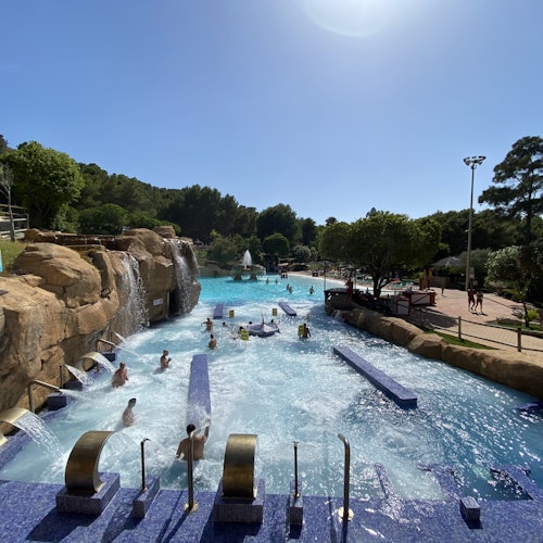 Personas disfrutando de una gran piscina al aire libre con chorros de agua, rodeada de rocas, árboles y un cielo despejado arriba.