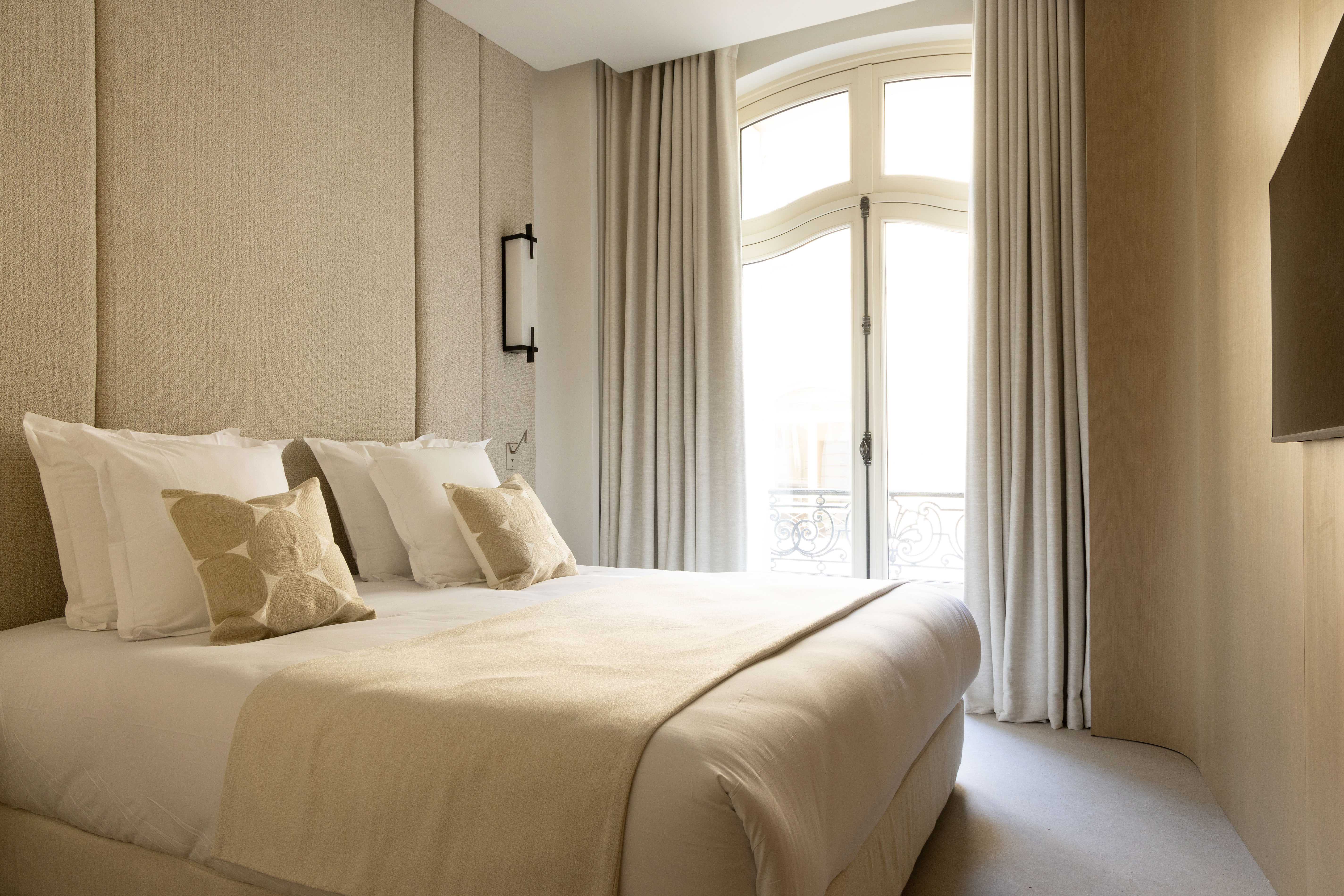 Chambre parentale tons creme avec tete de lit tissu, coussins beiges et fenetre cintree sur balcon parisien