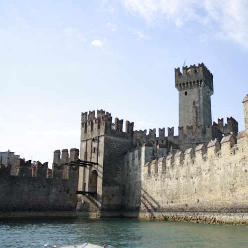 A medieval stone castle with turrets and battlements sits adjacent to a calm body of water under a partially cloudy sky.