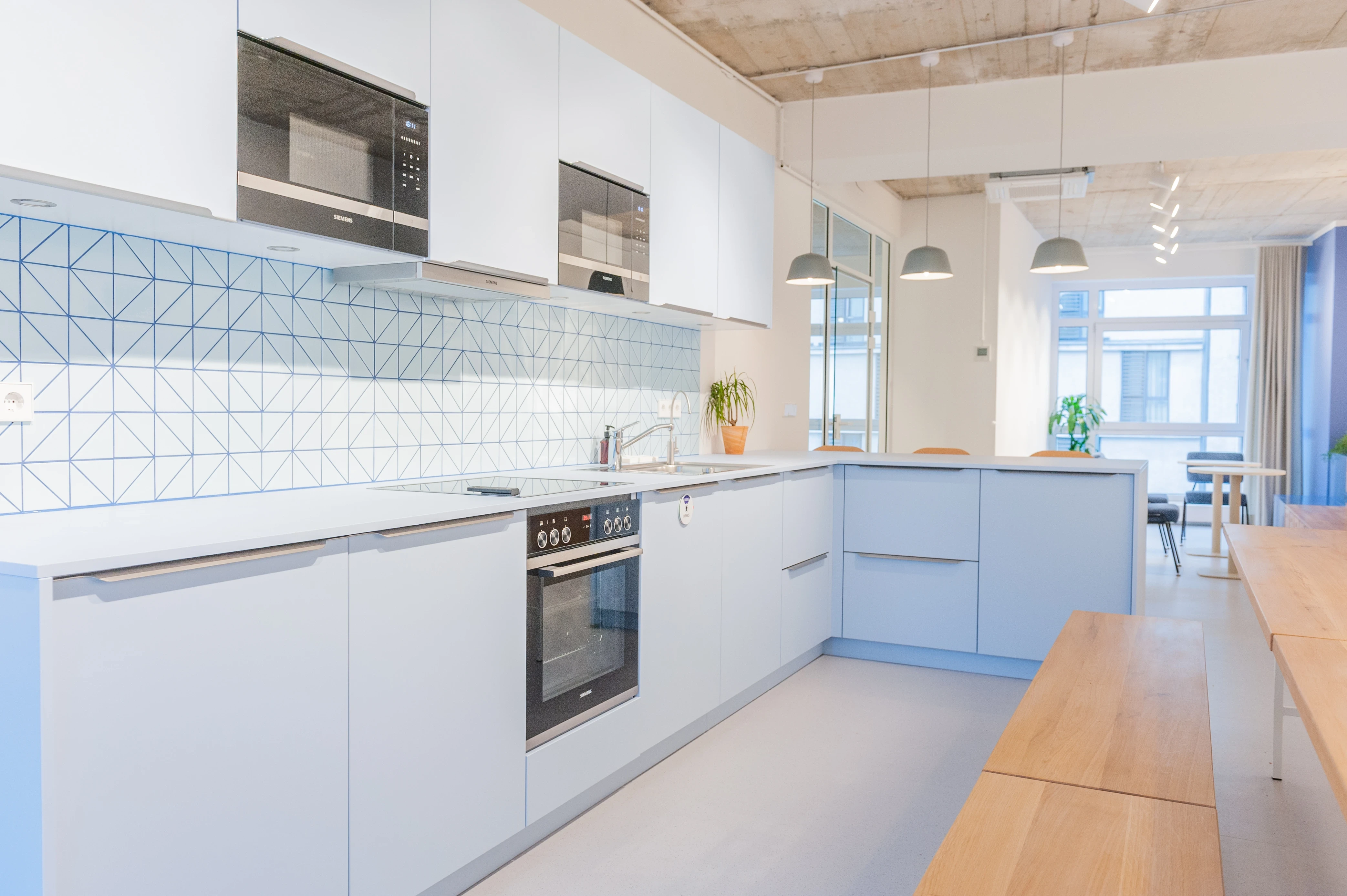 Kitchen and dining area at ZENLOOP with pale blue cabinets, geometric tiled splashback, grey pendant lights, a long wooden table with benches and a window-lit dining space.