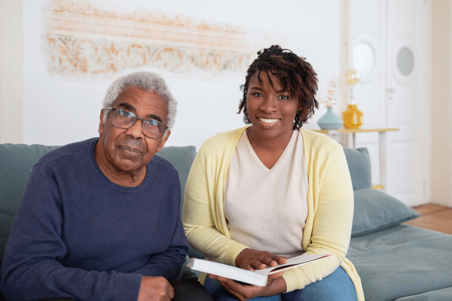 An older man sits with his daughter on the sofa at home, a book open between them.