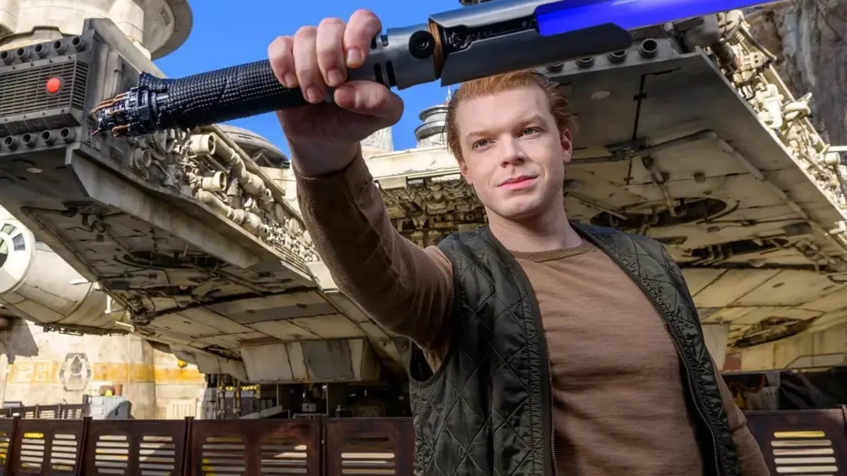 Cameron Monaghan holding a Disney replica of Cal Kestis's lightsaber in front of the Millennium Falcon at Disney's Galaxy's Edge