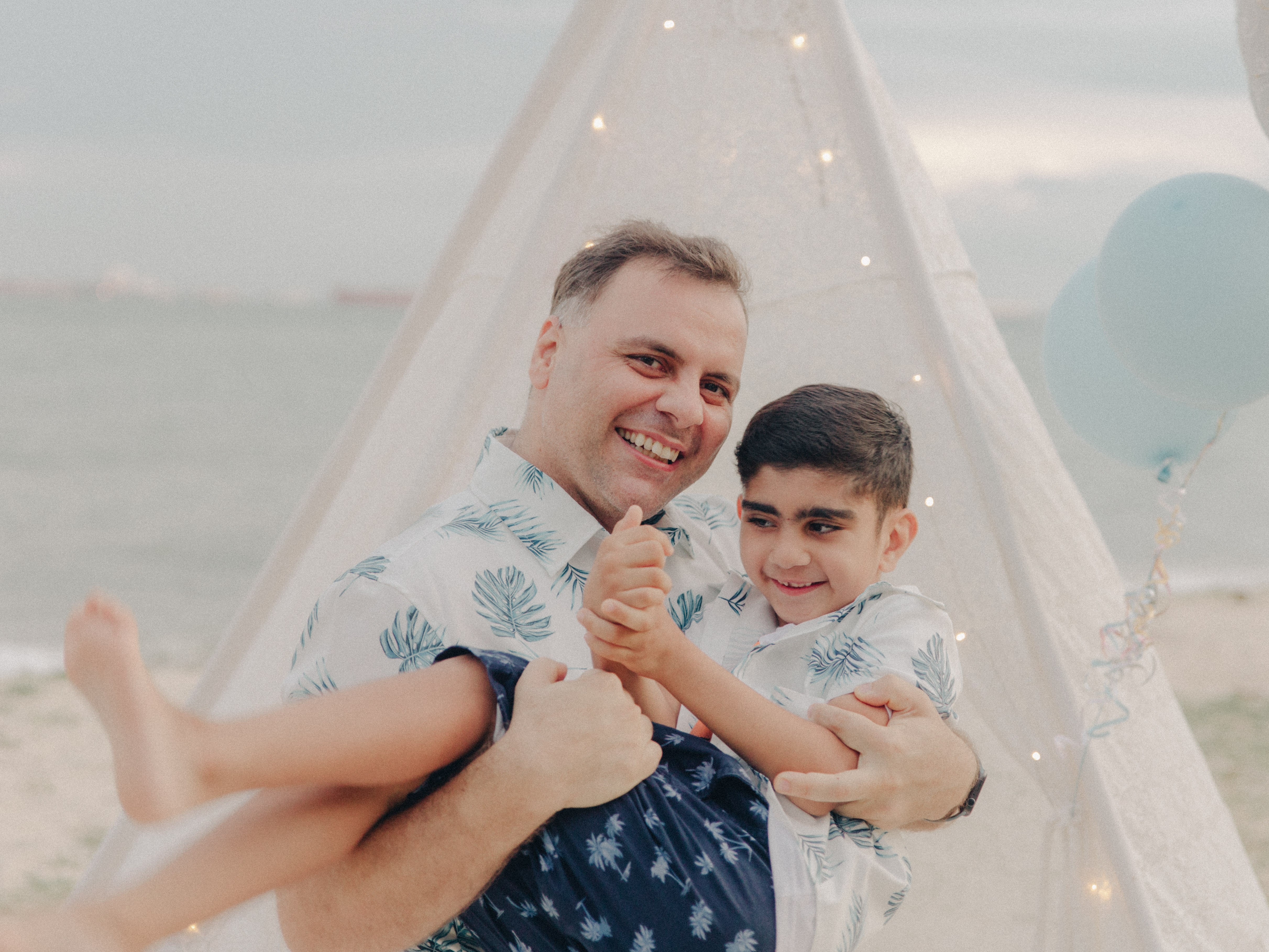 Father lifting his toddler son into the air by the beach, silhouette against the neutral sky.