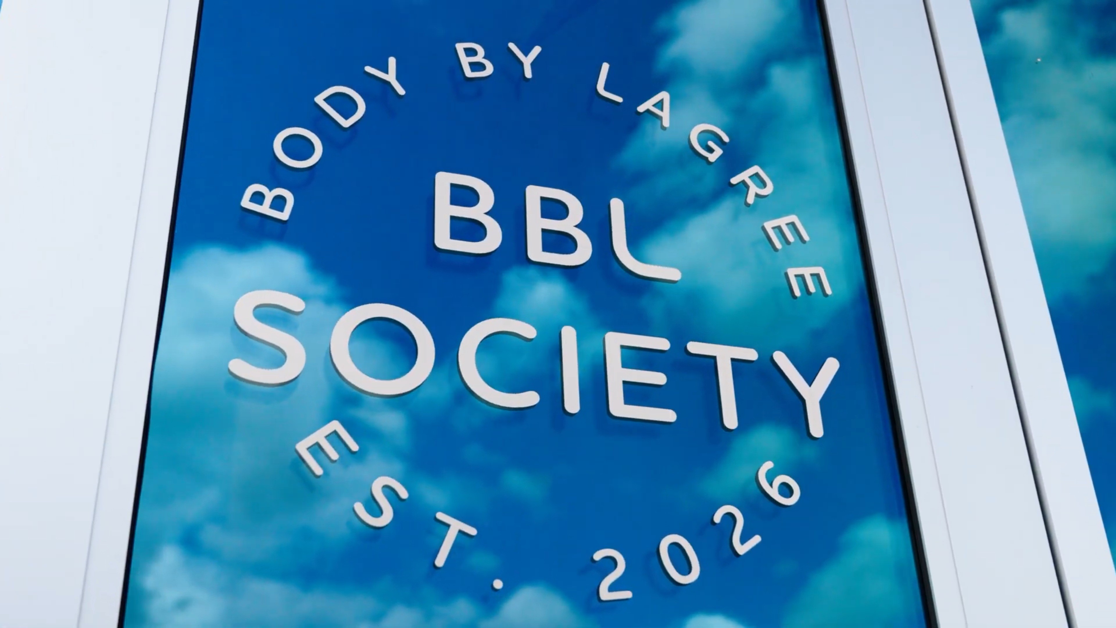 The BBL Society window sign — Body By Lagree, Est. 2026 — reflecting a blue Texas sky