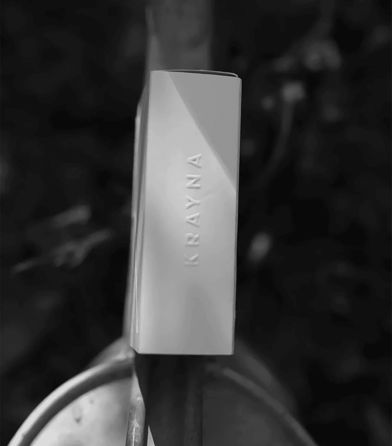 A black-and-white photograph of a Krayna product box balanced on a weathered outdoor surface. The contrast between the clean packaging design and the natural background highlights the brand's plant-based, organic roots.