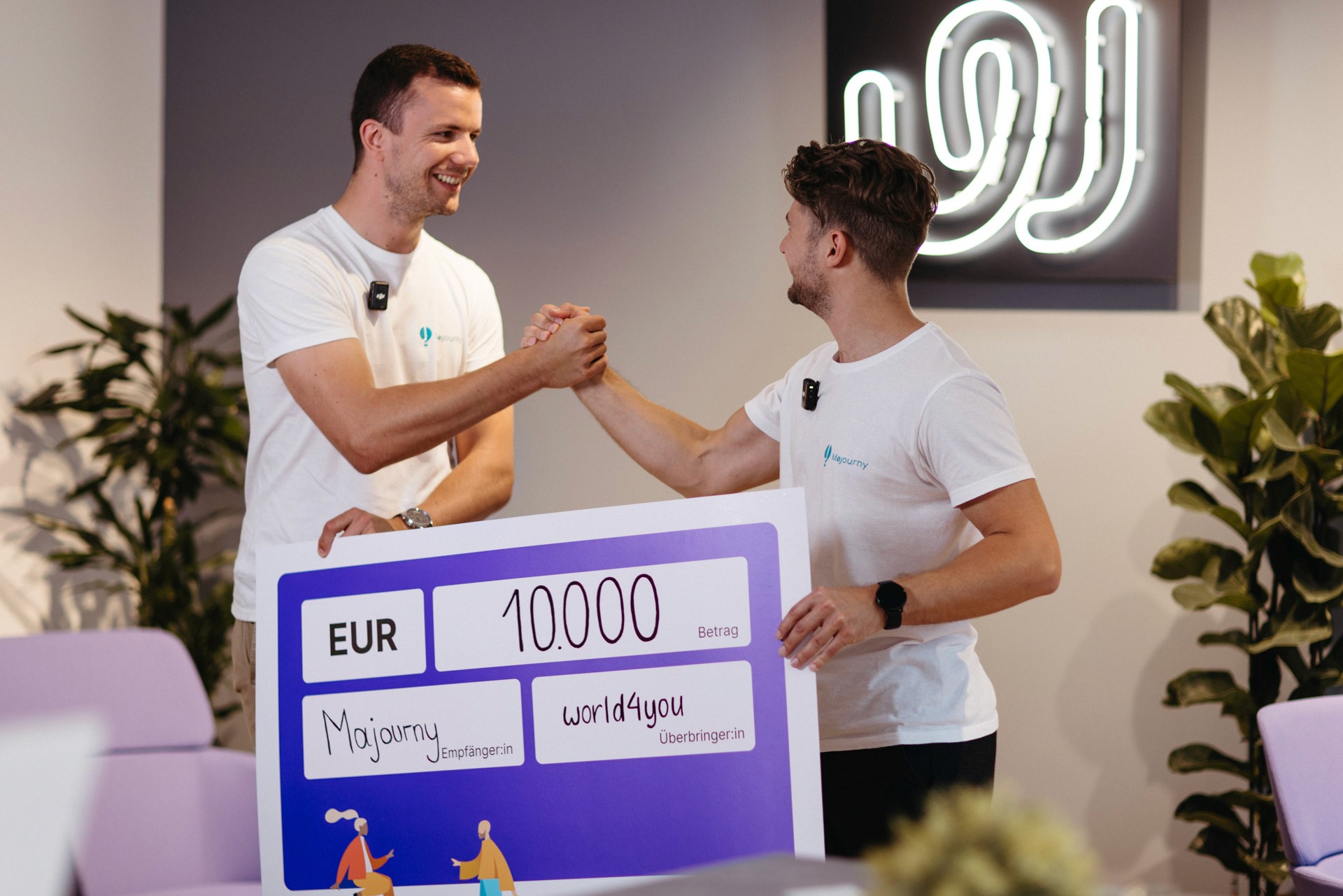 Two people in white shirts shake hands while holding a large check for 10,000 EUR in an indoor setting with plants and a bright sign in the background.