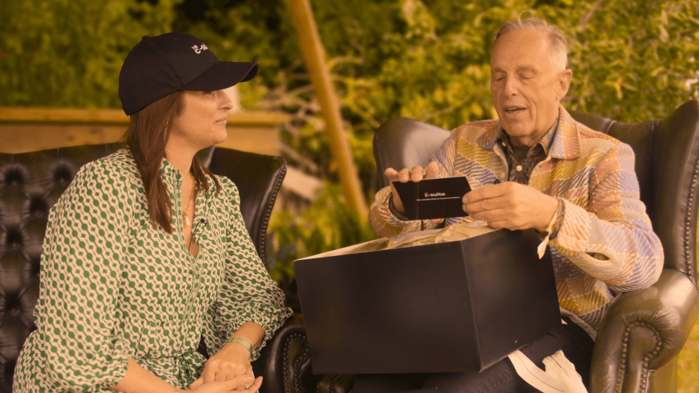 Young person with short, tousled blonde hair wearing an oversized, vibrant orange turtleneck sweater against a soft grey background, conveying a cool, modern look.