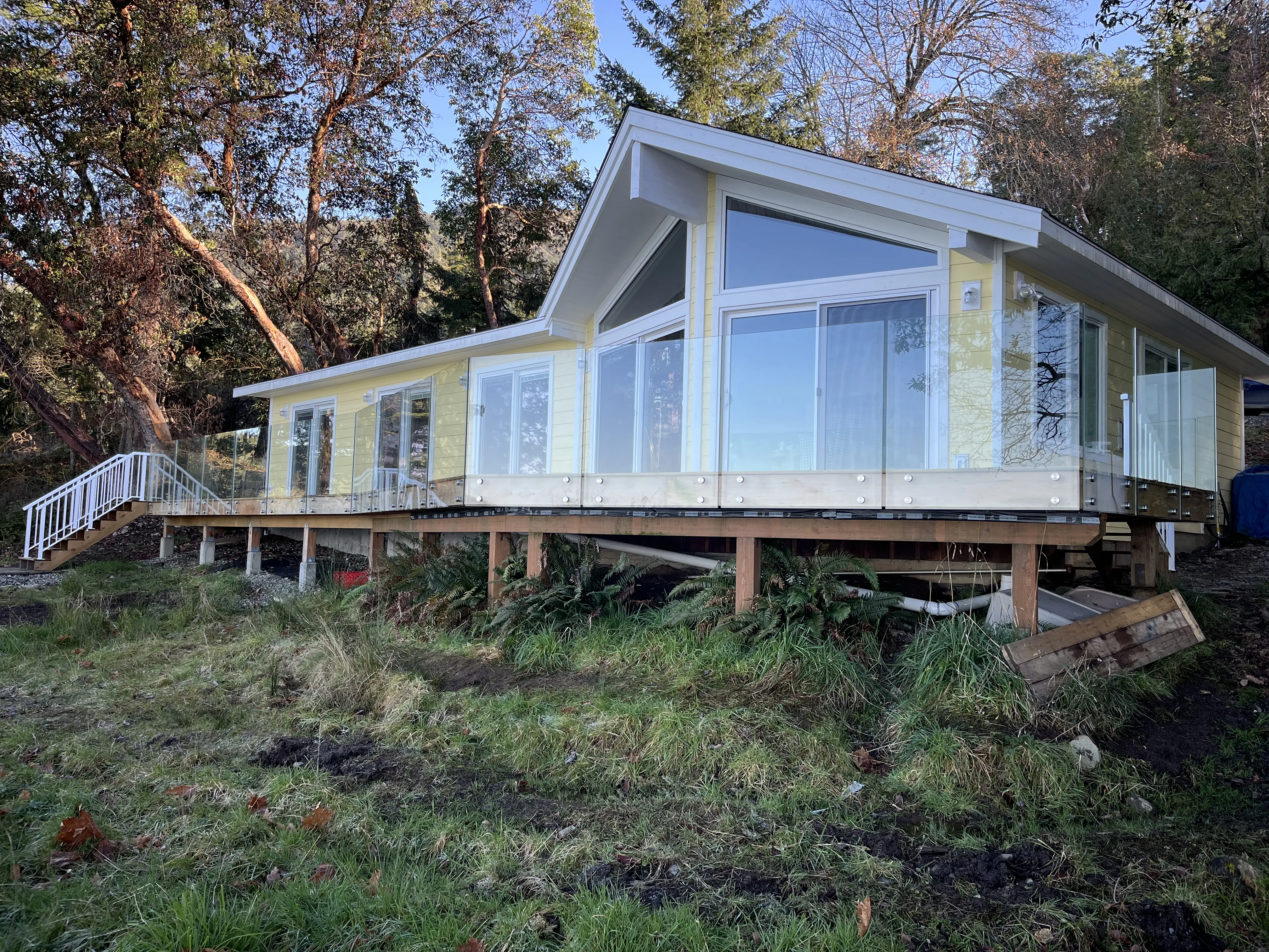 Single storey rancher with glass railings wrapping a raised wood deck on a treed coastal lot