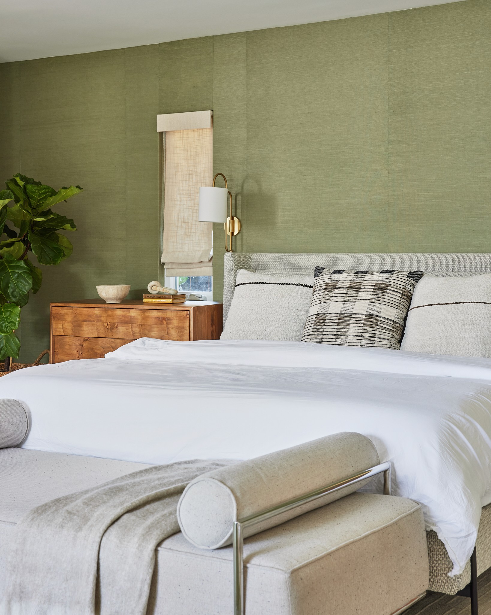 Master bedroom with green textured wallpaper, wall mounted reading light, tan pull down blinds, woven wood side dresser, tan upholstered bed with a bench and table at the foot, patterned pillows and throw, and teal rug by Jubilee Interiors in Los Angeles, California