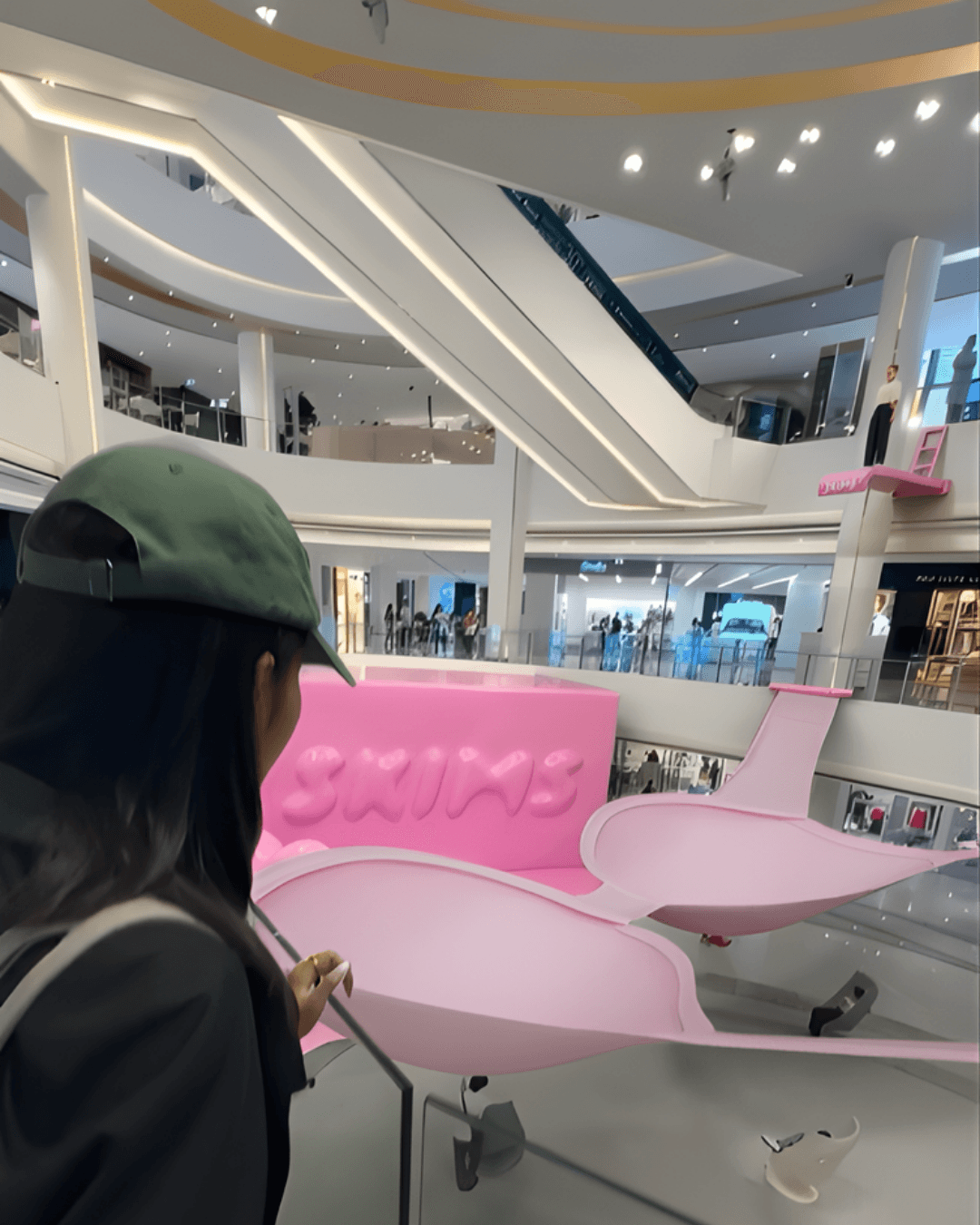 A mixed-reality mockup features a woman in a green hat looking over a railing at the giant pink installation