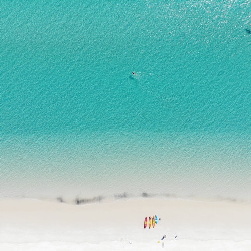 Aerial view of a turquoise ocean with a swimmer and four kayaks on a sandy beach.