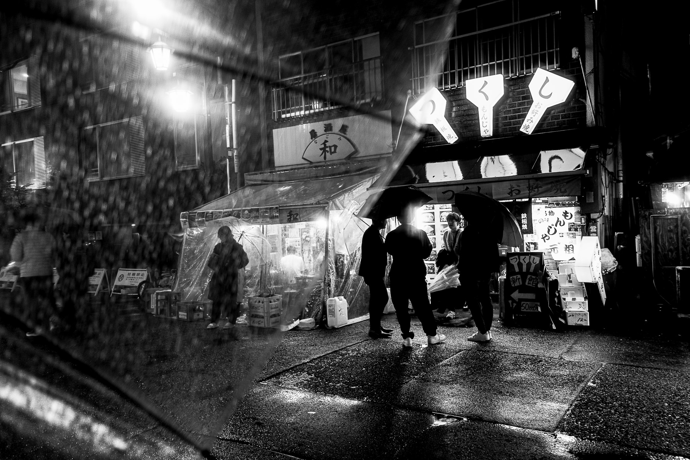 Black and white photograph of people walking with umbrellas on a rainy street at night, street photography by Richard Peterson.