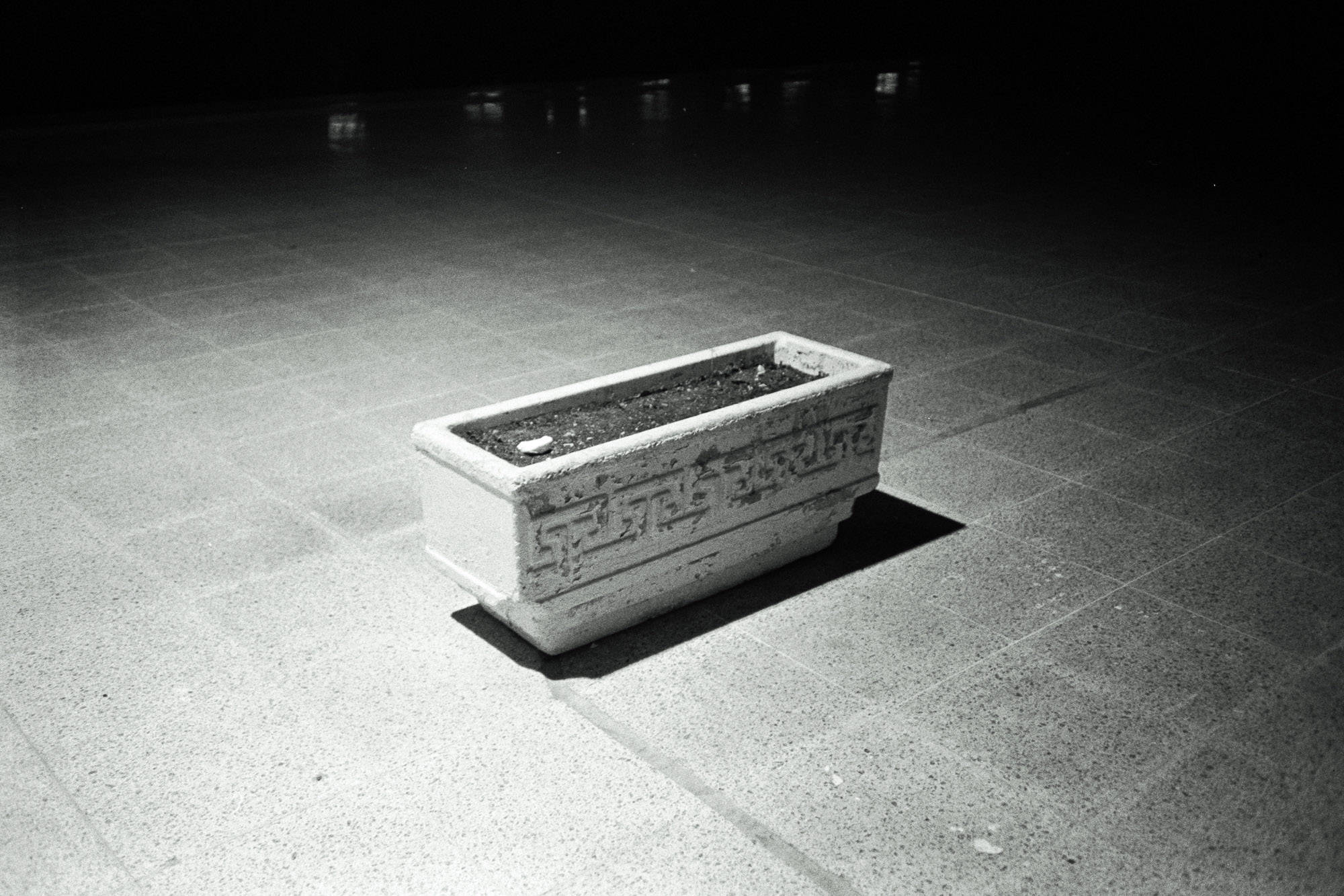 A rectangular stone planter sits empty on a tiled floor, illuminated by soft overhead lighting in a dimly lit room.