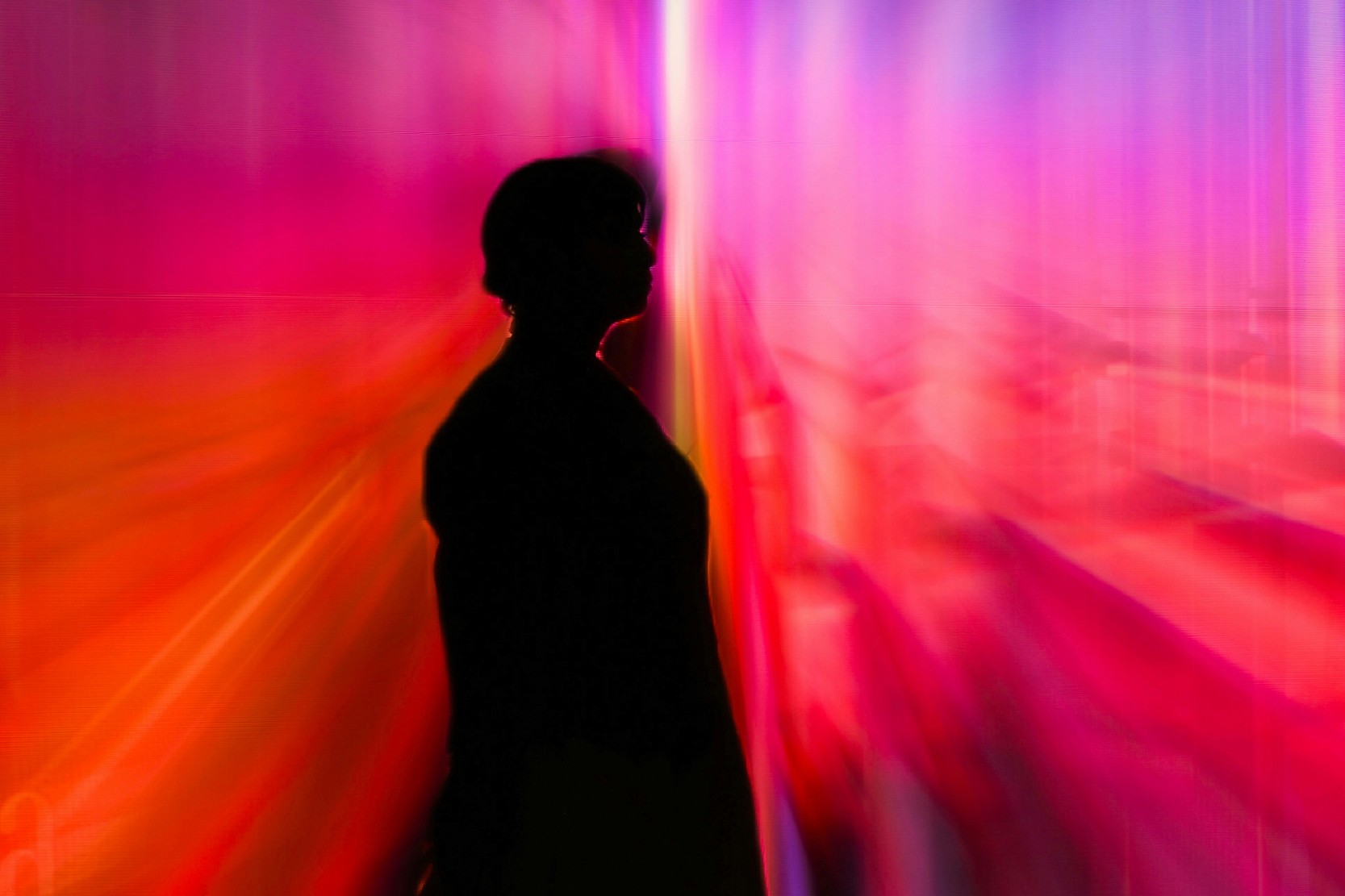Silhouette of a person in profile against a wall lit with streaking red, pink, and purple light.