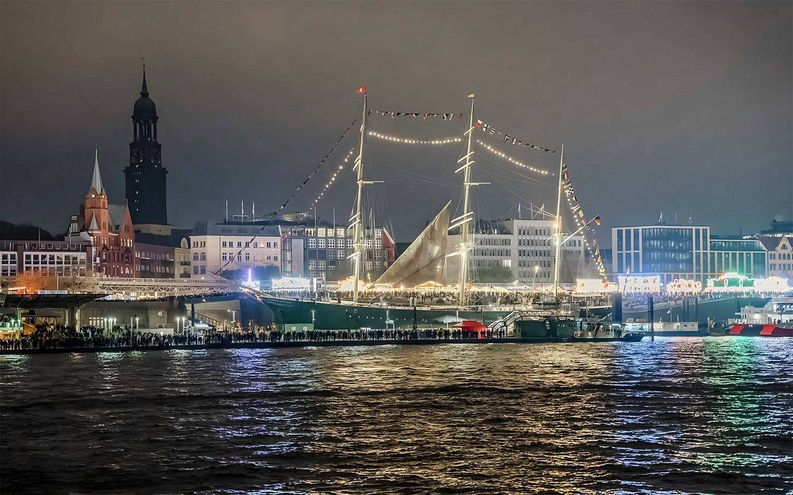 夜间灯光巡游期间，大港在夜色中被灯光照亮，周围有高大的船只与城市天际线。