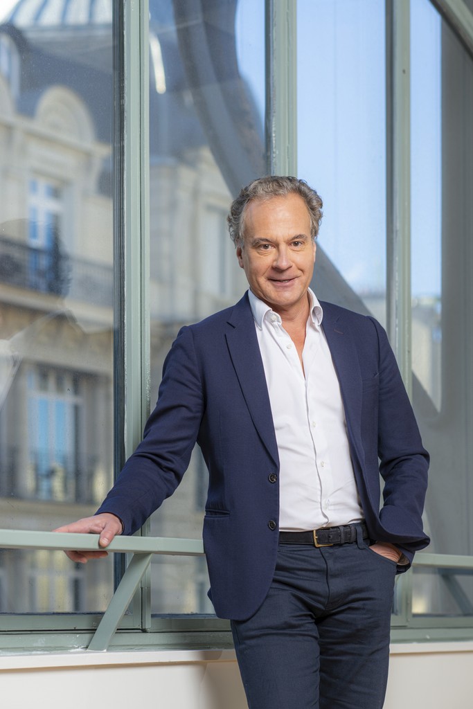 A man in a brown suit and cream silk scarf poses against a neutral background, exuding a composed and sophisticated demeanor.