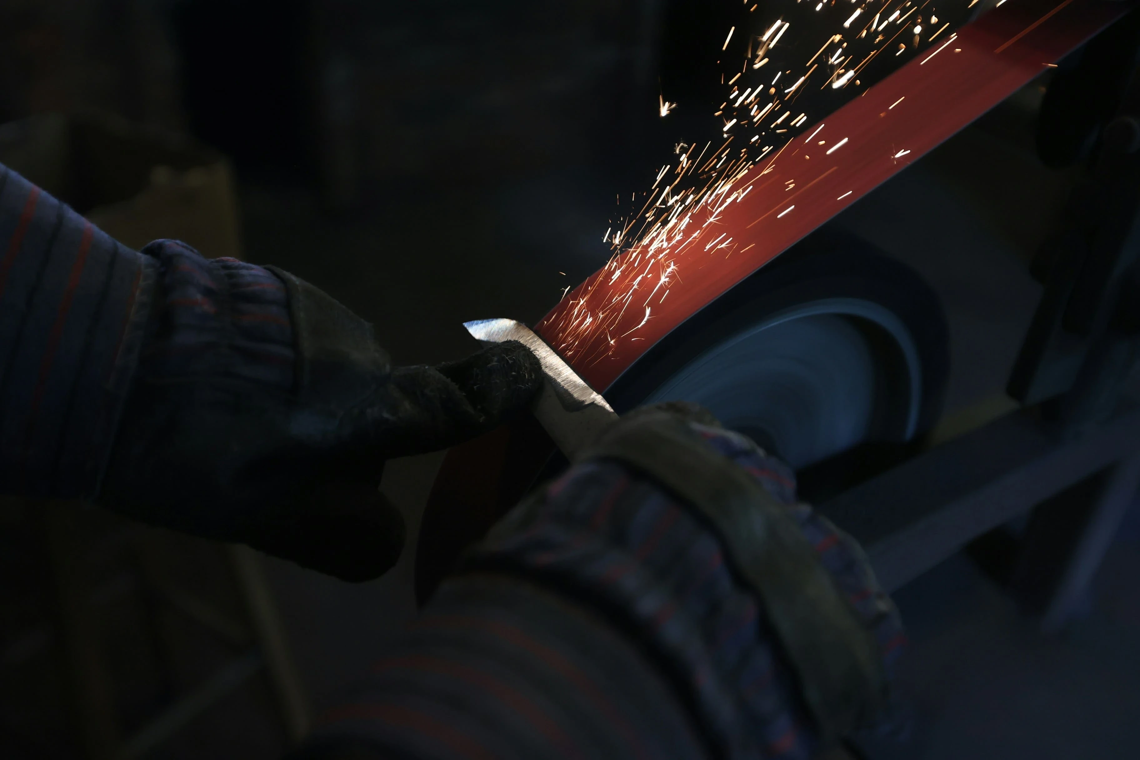 Close-up of a worker sharpening a metal blade on a grinding wheel, with sparks flying in an industrial workshop setting.