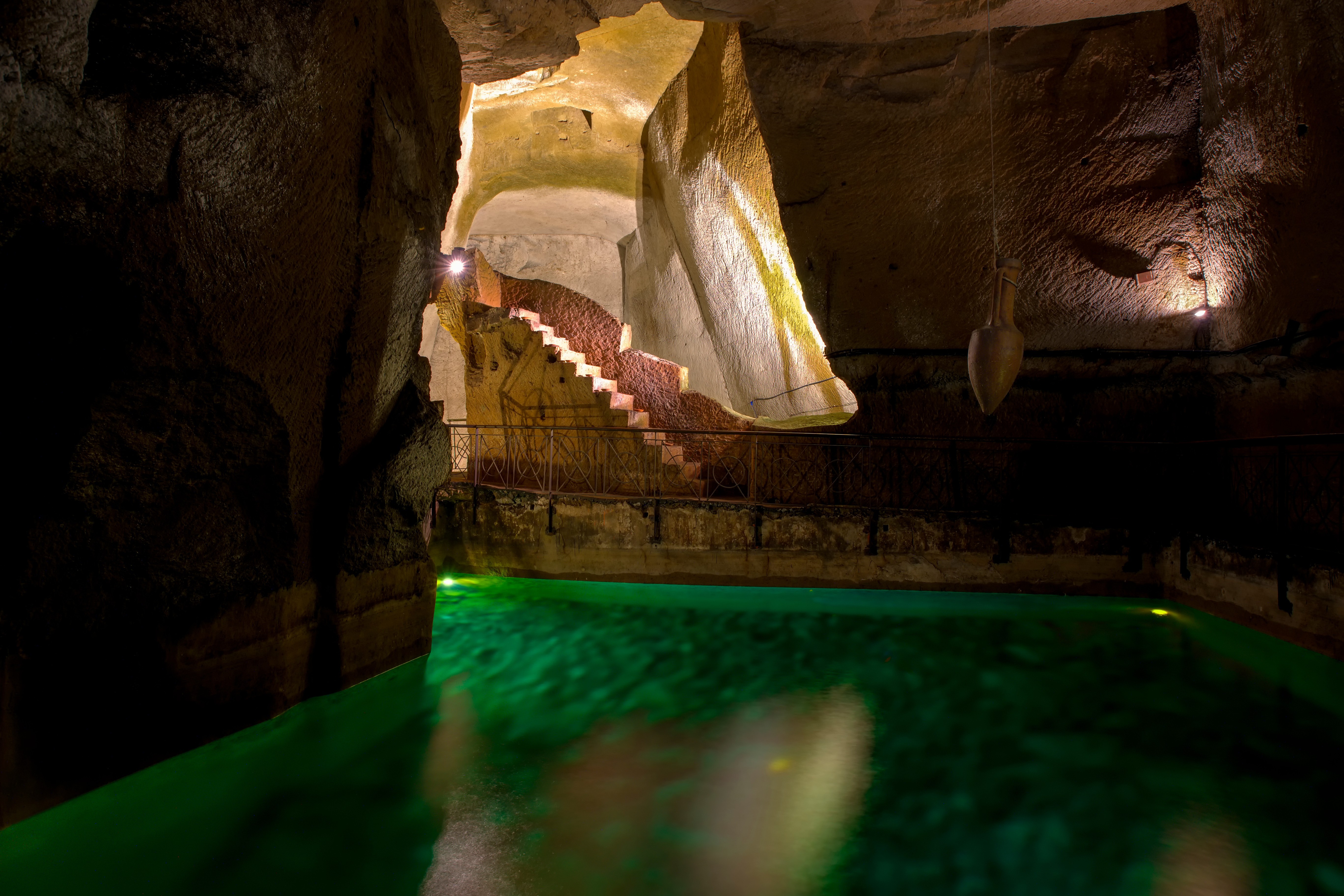 An ancient underground cavern in Naples, Italy, with a glowing green pool of water, rocky walls, and an illuminated stone staircase