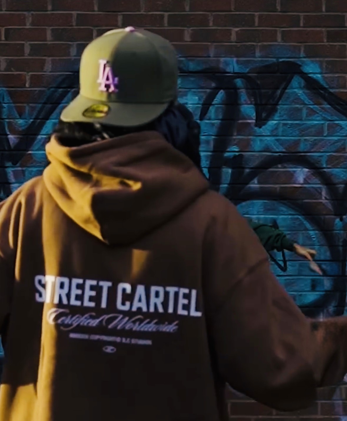 Streetwear photo of a model wearing a mustard Street Cartel hoodie against a graffiti wall.