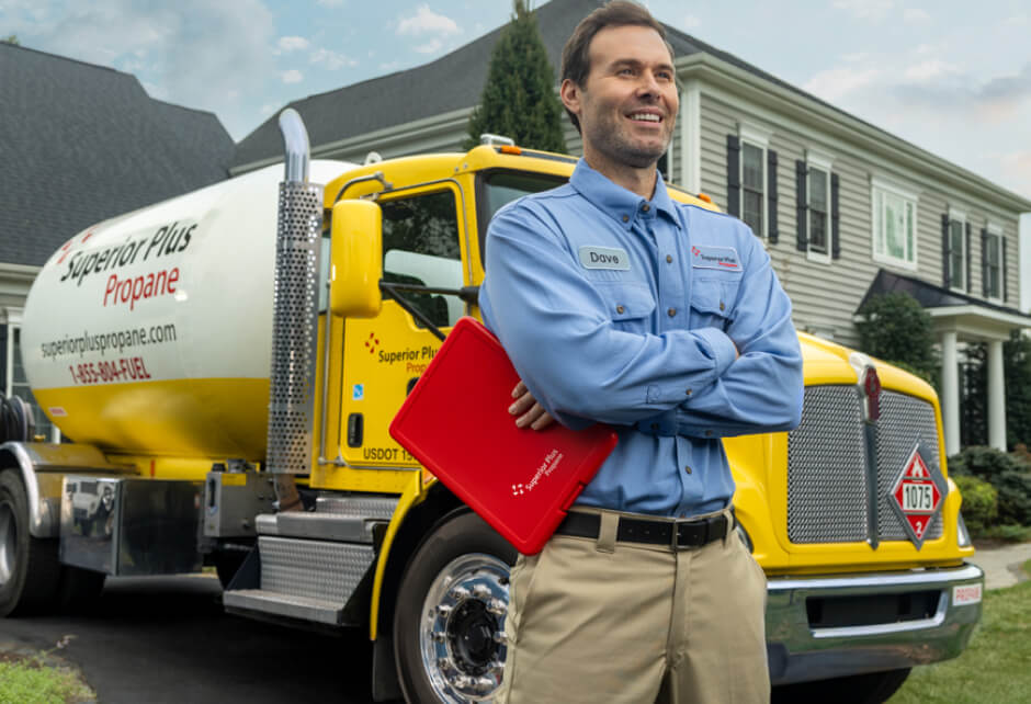 A man in a blue uniform stands in front of a yellow propane delivery truck, holding a red folder, outside a suburban house.