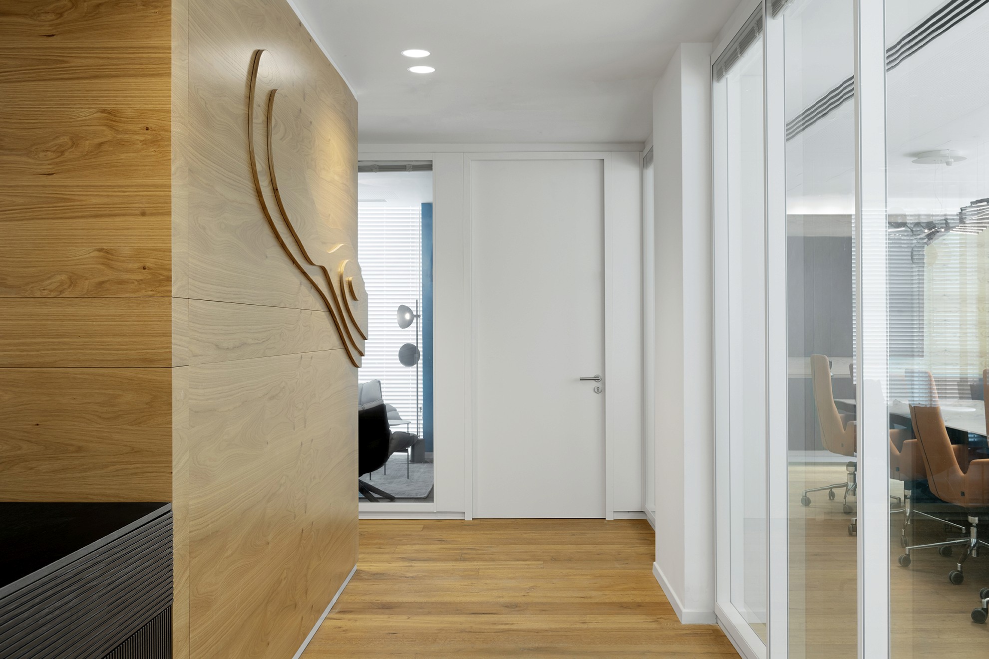 זרימה בחלל: מבט לאורך המסדרון המציג את קיר הטופוגרפיה מעץ המלווה את המעבר, לצד מחיצות זכוכית שקופות למשרדים.