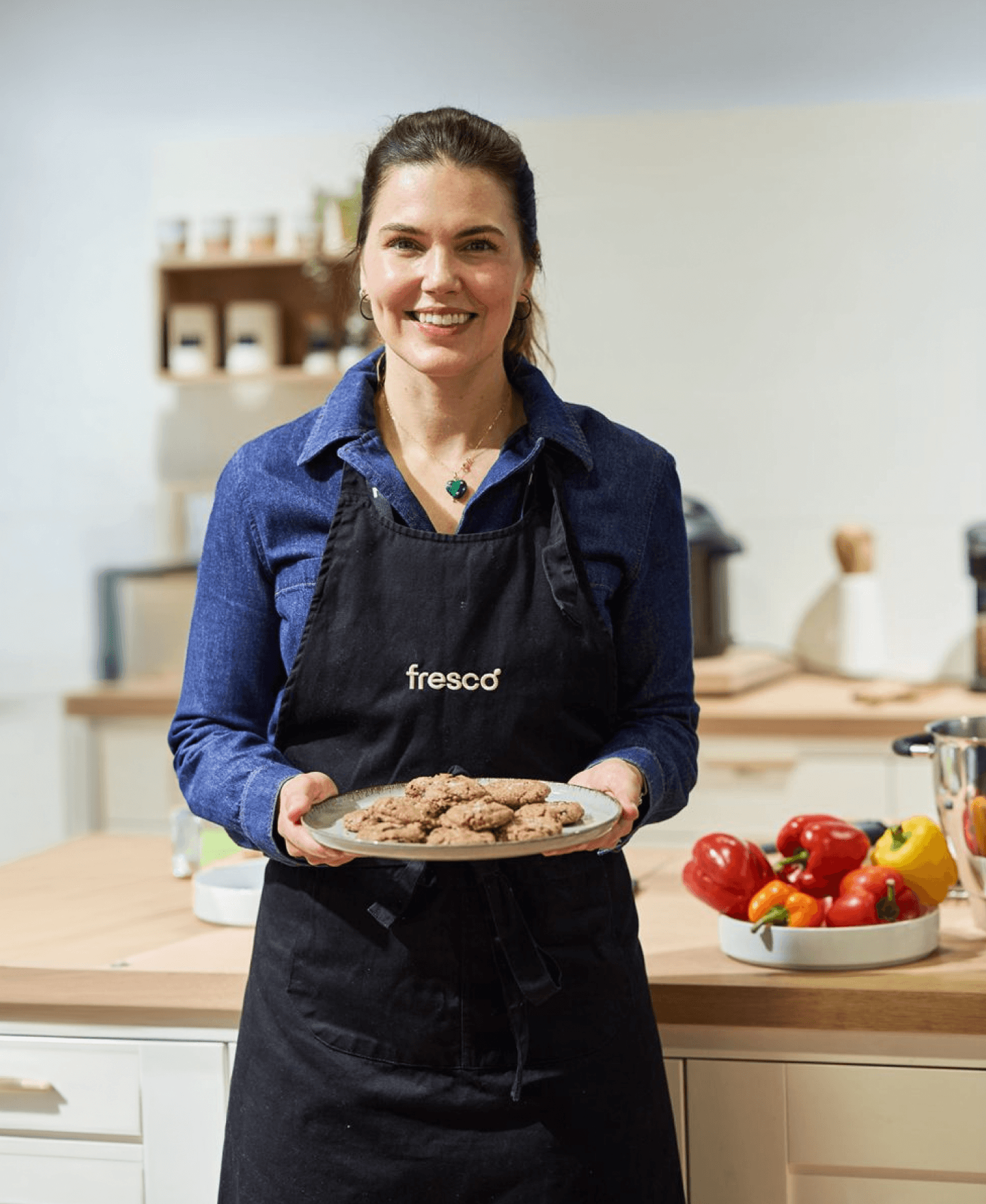 Woman wearing a Fresco apron smiles while holding a plate of freshly baked cookies in a bright kitchen