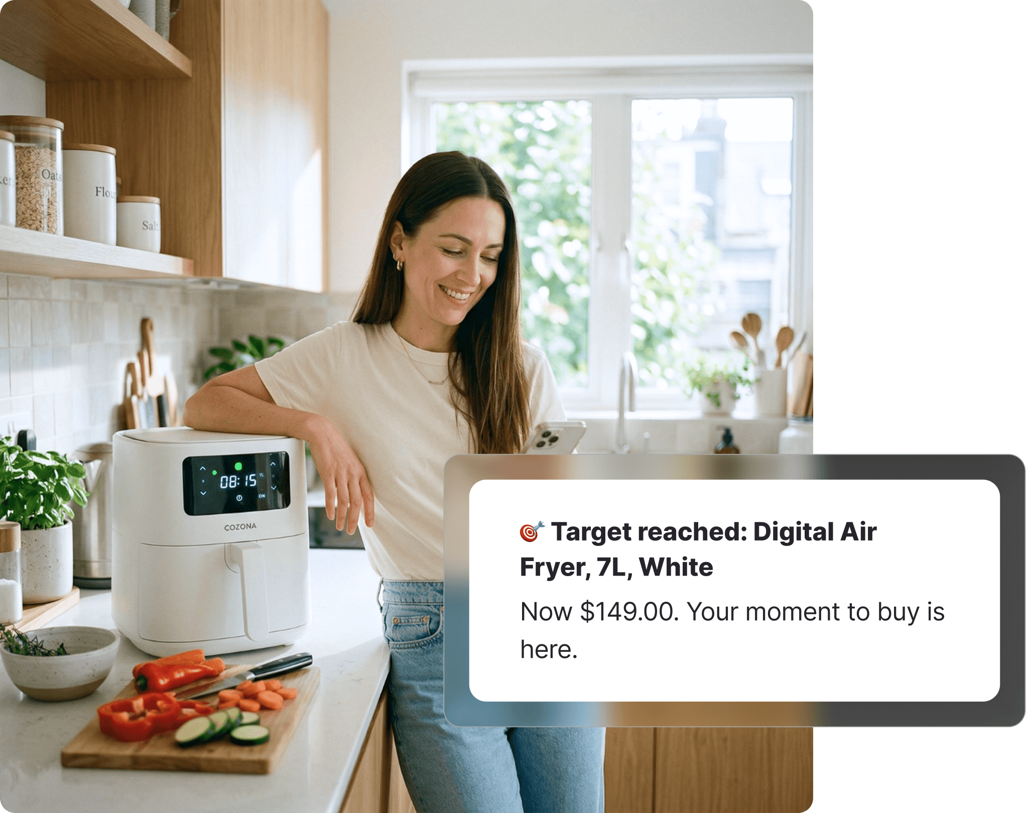 Woman in a kitchen smiling at her phone next to a white air fryer, with fresh vegetables on a chopping board beside her, receiving a Harvey Norman target price notification on Whisprice.