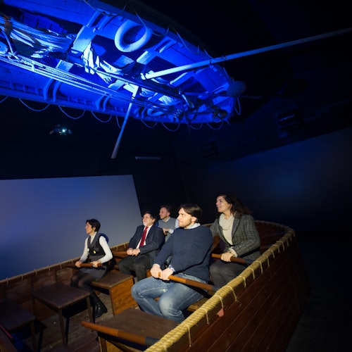 青い照明と天井の装飾が施された暗い部屋で、木製のボートのレプリカに座り、スクリーンを見つめる人々。