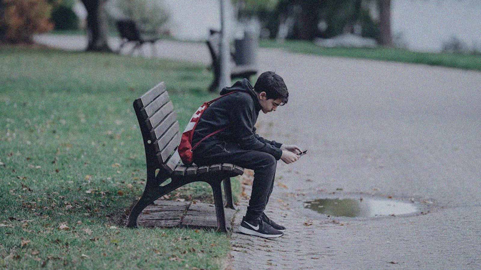 Imagen de un niño sentado en una banca mirando su celular en la columna de opinión "El desafío detrás de la violencia" de IdeaPaís.