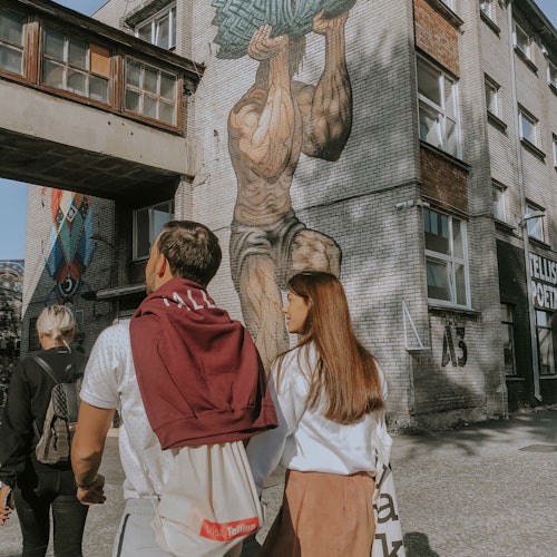 Három ember sétál egy épület mellett, amelyen egy freskó látható, amely egy izmos alakot ábrázol, amint egy nagy terhet tart fel.